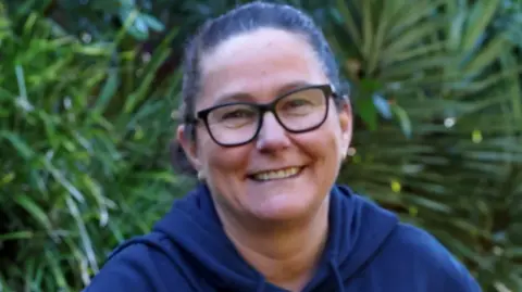 Supplied: Elizabeth Rowley A middle-aged woman wearing glasses and with her dark hair in a pony tail smiles at the camera
