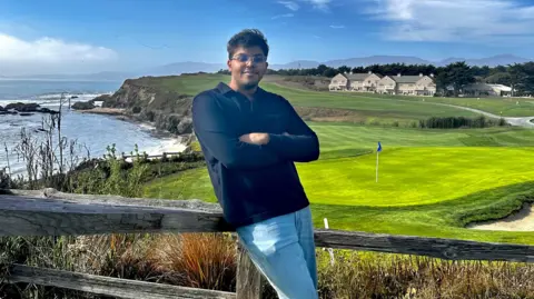 Ayush Deb Ayush Deb poses for a picture with his arms crossed in front of a golf course by the sea.