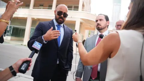 PA Joseph McBride, bald and with a beard and mustache and wearing sunglasses and a blue suit, shirt and tie, and Thomas Maniotis, with short cropped brown haior and a beard and mustache, with a grey suit and red tie, speak into microphones surrounded by media