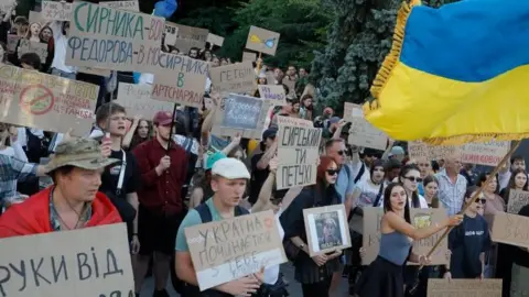 EPA/Shutterstock Dozens of people, mostly young, holding cardboard signs in a protest, as one woman waves a large Ukrainian flag