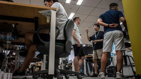 Getty Images Four men wearing shorts in the office in Tokyo, Japan. Three of them standing up, one of them seated and with his elbows resting on his table.