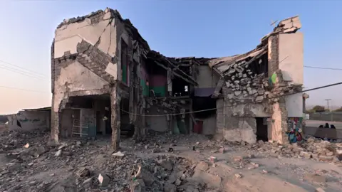 JACK GARLAND / BBC The section that remains of Shajareh Tayyebeh Primary School in Minab. It is a severely damaged two-storey building, with sections of wall missing and chunks of concrete hanging off it. It is surrounded by a flat, rubble-strewn area.
