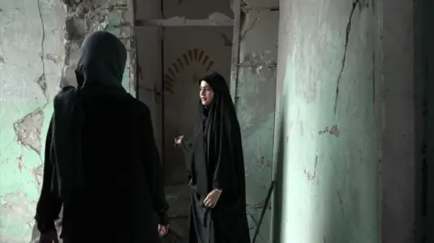 JACK GARLAND / BBC Amaineh Nademi walking along a remaining corridor of the Shajareh Tayyebeh Primary School in Minab. She is wearing a black chador and showing BBC reporter Nawal Al-Maghafi the damage to the school. The concrete walls are cracked, and have previously been painted pastel green.