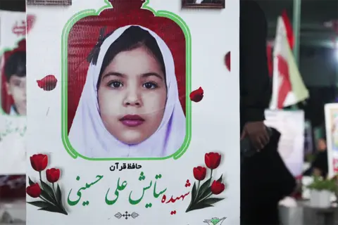 JACK GARLAND / BBC A photo of nine-year-old Setayesh displayed on her grave. She is wearing a white veil and looking at the camera. The image is surrounded by a white frame decorated with red flowers. In the background other graves and an Iranian flag can be seen. 