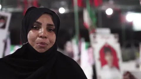 JACK GARLAND / BBC Masoumeh Mehran Shekhabadi pictured in a black chador with a sad expression. Behind her in soft focus are images of children on graves in the cemetery