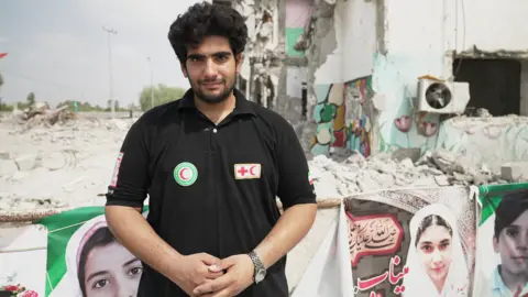 JACK GARLAND / BBC Red Crescent rescue worker Reza Besharati, pictured in a black T-shirt with Red Crescent badges on it. He is standing in front of banners showing the faces of people killed in the attack on the school, with the damaged buildings behind him.