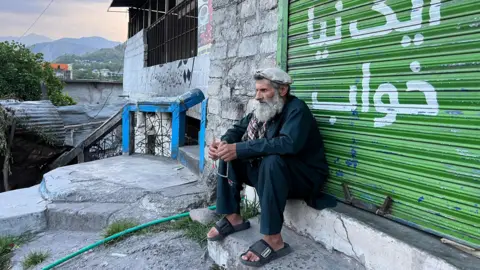 Naseer Chaudhry A man with a beard sits on a step in front of a bright green roller door. He is wearing a hat and flip-flops, and holding prayer beads in his fingers while staring forward pensively