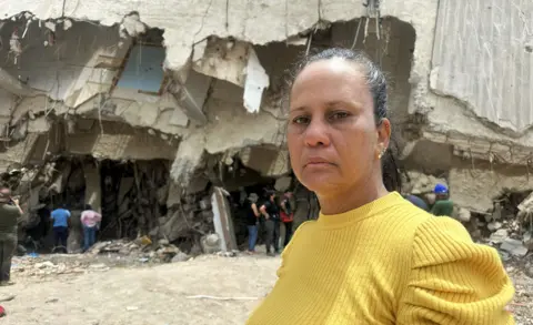 Laura Barrios stands in front of a building as she searches for the remains of her brother-in-law. She is wearing a yellow sweater, her dark hair is pulled back.