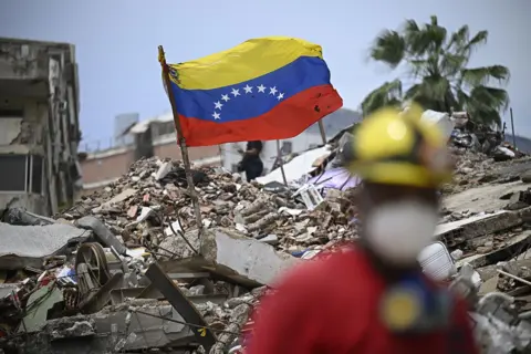 Federico Parra / AFP via Getty Images A Venezuelan flag is pictured on top of a pile of rubble with a palm tree in the background against a blue sky, while a rescue worker walks in the foreground with bokeh, in Caraballeda, Venezuela on 20 July.