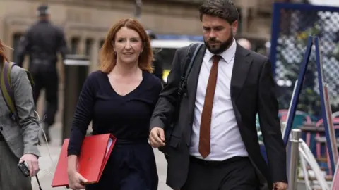 PA Media Louise Haigh is walking down the street next to an aide. She is carrying a  large, scarlet government folder and wears a smart black top and black trousers. Her hair is ginger and shoulder length, and she wears it loose, along with a pair of small earrings.