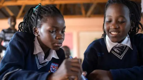 AFP via Getty Images Two 13-year-old girls, who are members of the Ugandan dance group the Ghetto Kids, attend school in Kampala on 20 Apri 2023. They are looking down at a book and wearing navy school jumpers and white shirts.
