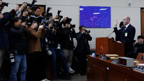 SeongJoon Cho/Bloomberg via Getty Images Harold Rogers from Coupang is seen holding his hand up to take an oath during a hearing into a data breach while a bank of photographers take images.