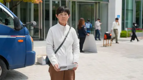 BBC/Hosu Lee A middle-aged woman with short, dark hair wearing a white fleece stands outside a building