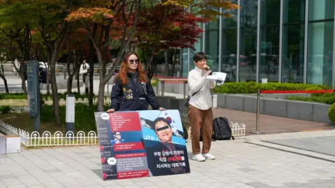 BBC/Hosu Lee Two women stand outside a building in Korea. One talks into a microphone, the other holds a large poster in protest.