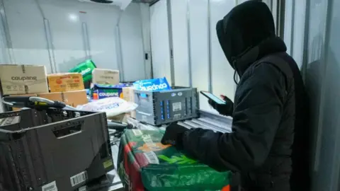 BBC/Hosu Lee A person wearing a black hoodie and holding a phone is seen from behind as they look into the back of a well-lit truck, with various containers and packages in it.