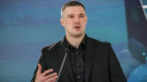 Reuters A man in a black jacket and black shirt talks in a microphone with a blue backdrop. His hair is close-cropped and he looks passionate.