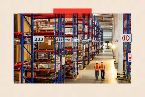 Getty Images Two people in white hats and orange vests walk across a warehouse