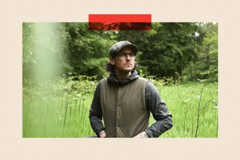 A photograph of a man in a field wearing glasses, a flat cap and a khaki jacket