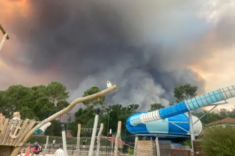 Andrea Gretton A huge cloud of dark grey smoke approaches a campsite where children's play equipment can be seen in the foreground.