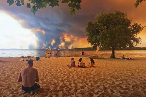 AFP via Getty Images Beach goers rest on the beach of Moutchic on the shore of Lake Lacanau, southwestern France on July 24 as clouds of smoke rise into the sky from the Gironde wildfire. Huge smoke clouds and an orange sky are seen in the distance across the water, while people sit on the beach in the foreground.