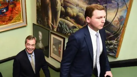 PA Media George Cottrell, in suit and tie, walks up a staircase past a wall decorated with pictures with Nigel Farage, also in suit and tie, following     