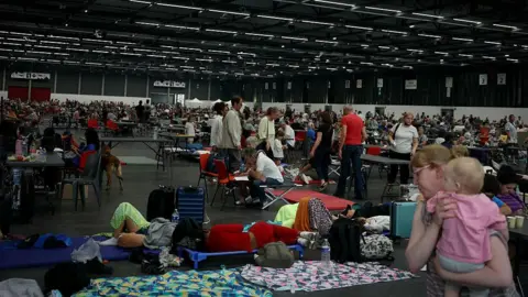 AFP via Getty Images A number of people shelter at an exhibition centre - some people sleep on mats on the floor, while a woman carries a baby on the bottom right of the photo