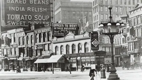 Getty Images Historic black and white photo showing an ornate streetlamp in front of buildings and old billboards