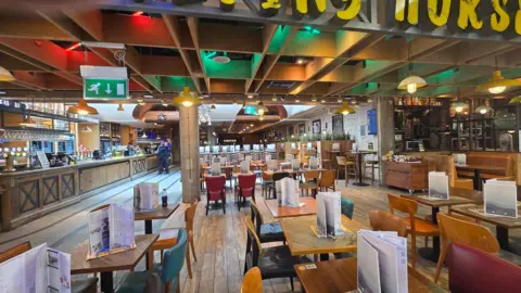 Roger Wood Interior of a large airport restaurant showing tables and chairs, menu displays, a bar counter and overhead lighting. Only a small number of people are visible near the bar area while much of the seating is empty.