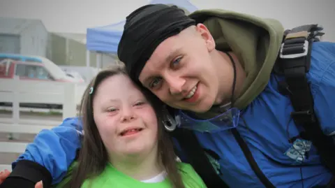 Down’s Syndrome Association Gracie poses alongside Aitch, who has his arm around her. Gracie is wearing a bright green t-shirt. Aitch is wearing a blue skydiving jumpsuit. 
