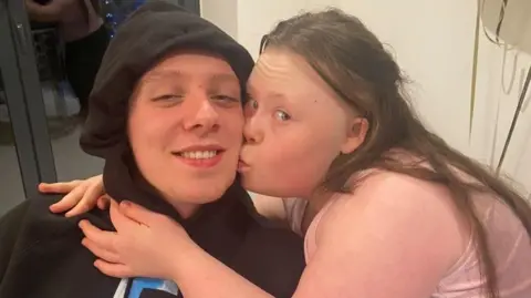 Handout Aitch posts with his sister Gracie, who is kissing him on the cheek. Aitch is wearing black hoodie with the hood up. Gracie is wearing a pink top.  