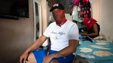 Lee Durant/BBC A man with a pistol sits on a bed. He's wearing blue shorts, a white Tommy Hilfiger t-shirt, a black cap and a red balaclava obscuring his face. 