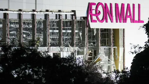 AFP via Getty Images A damaged wall is seen on the side of Aeon Mall Kumamoto. The words Aeon Mall are seen in bright pink letters.