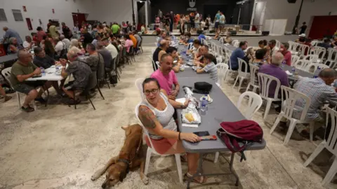 Reuters Wide shot of a shelter in Spain with three long plastic tables and plastic garden chairs around them. People evacuated from their homes are sitting at the tables eating and talking