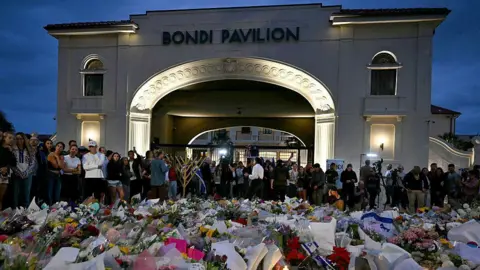 AFP via Getty Images A large arched building in the background says "Bondi Pavilion". In the foreground, a large collection of tributes - made of of flowers, Israeli flags and other items - is laid on the floor in front of a crowd of people who have gathered. 