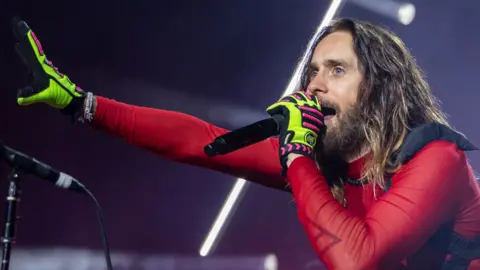 Getty Images Jared Leto stands under concert lighting, singing into a handheld microphone. He has long hair and a beard and is dressed in a red, sheer and fitted top with neon green and pink gloves. One arm is stretched outward toward the audience while the other holds the microphone close to his mouth. Bright white stage lights form diagonal lines in the background.


CLARKSTON, MICHIGAN - AUGUST 06: Actor/Singer Jared Leto of Thirty Seconds To Mars performs during their Seasons World Tour 2024 at Pine Knob Music Theatre on August 06, 2024 in Clarkston, Michigan. (Photo by Scott Legato/Getty Images)