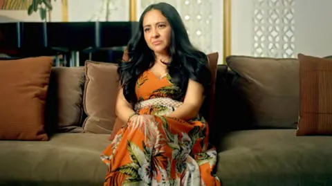 Isabel sits in the middle of a velvet green sofa with brown pillows behind her. She has a serious expression and long black wavy hair. She is wearing an orange dress with a floral pattern on it.  