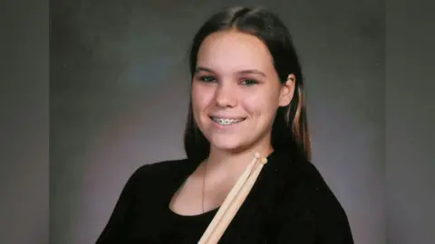 Taylor when she was 14 standing against a soft gray studio backdrop for a formal portrait. She is wearing a plain black short-sleeved top and holding two wooden drumsticks upright across the chest. Long dark hair falls past the shoulders, and she is smiling, with metal braces visible on her teeth.