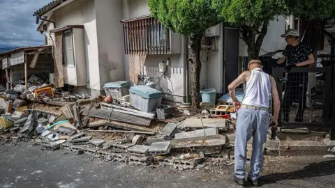 Getty Images A man in a singlet and blue jeans surveys the debris outside a house. Another person in a cap and gloves looks on from behind a fence