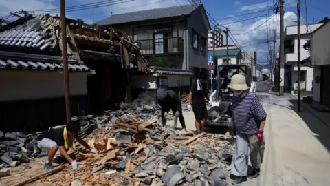 Reuters Four people picking up debris from a partially collapsed temple, with a small digger behind cleaning the rubble