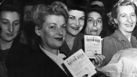 Getty Images This black-and-white photograph shows a group of people standing closely together in what appears to be an indoor public setting. Several individuals in the foreground are holding up documents clearly labelled “RATION BOOK”. The crowd fills most of the frame, and the image focuses on the ration books as key objects, suggesting a scene connected to food rationing or distribution. 