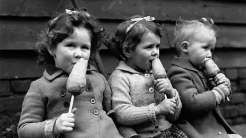 Getty Images This black-and-white photograph shows three young children sitting side by side outdoors in front of a wooden plank wall. Each child is holding and eating a root vegetable on a stick. The children are dressed in coats or cardigans suitable for cool weather, and two of them wear ribbon bows in their hair. 