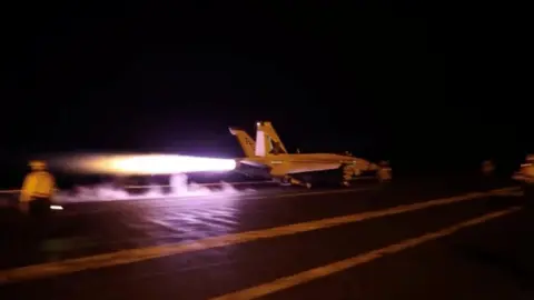 US Central Command A large flame blasts from the back of a fighter jet on a runway with an air traffic controller stood nearby