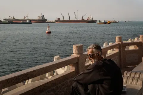 Getty Images A man with grey hair, black clothing and sunglasses looks on from a stone observation point in Oman at the Strait of Hormuz. In the distance are at least five boats and a buoy floating on the water.