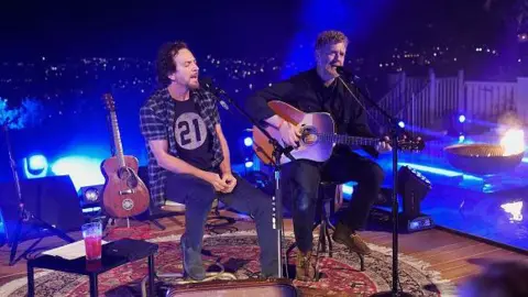Getty Images Eddie Vedder is wearing a checked shirt and black t-shirt and jeans and has dark hair and a goatee beard he is singing beside Glen Hansard who is playing guitar and singing.