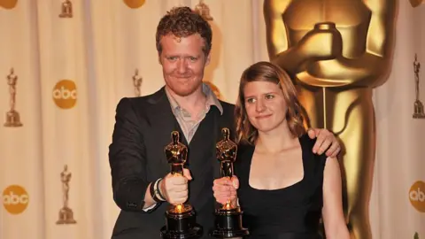 Getty Images Glen Hansard and Markéta Irglova hold their Oscars. Hansard has short red hair and stubble and is wearing a suit jacket and open-necked shirt. Irglova is wearing a black dress and has shoulder-length reddish/brown hair
