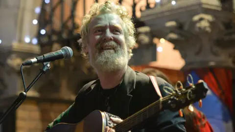 Nur Photo/Getty Images Glen Hansard smiling as he plays guitar outside a Dublin theatre on Christmas Eve night 2019.  He is a man with greying ginger hair and a matching beard.  He is wearing a dark green blazer over a dark t-shirt 