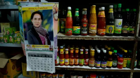 BBC/ Lulu Luo A calendar with the image of Aung San Suu Kyi hangs beside bottles of Burmese sauses and condiments in Mae Sot Thailand, which borders Myanmar
