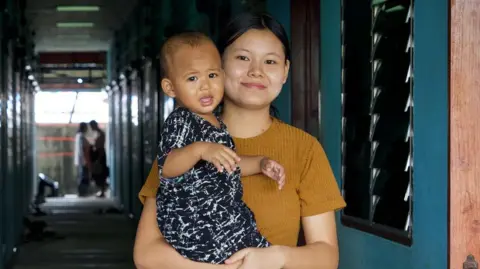 BBC/ Lulu Luo Young Burmese woman wearing a brown shirt, her hair tied, holding a baby wearing a black and white shirt and shorts, with an open door and a corridor in the background in Mae Sot, Thailand, that borders Myanmar