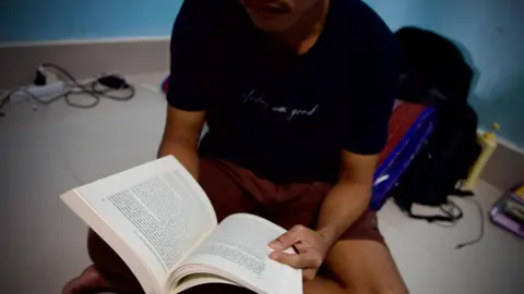 BBC/ Lulu Luo A Burmese man wearing a black t-shirt, sitting on the floor, with a book in his hand, in a room with blue walls.