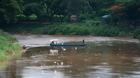 BBC/ Lulu Luo People ride a blue boat on a river with brown water, with thick foliage in the background in Mae Sot Thailand, which borders Myanmar